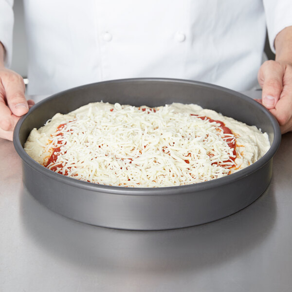 A person using an American Metalcraft Hard Coat Anodized Aluminum Cake Pan to prepare a pizza.