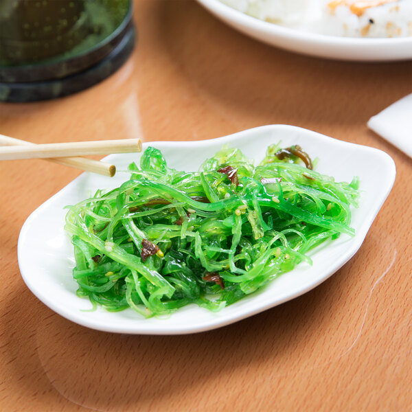 A close up of a bright white irregular oval porcelain dish with green seaweed salad on it with chopsticks.