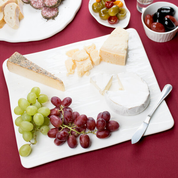 A white rectangular wood grain porcelain platter with cheese, grapes, and a round white cheese with a cut out slice.