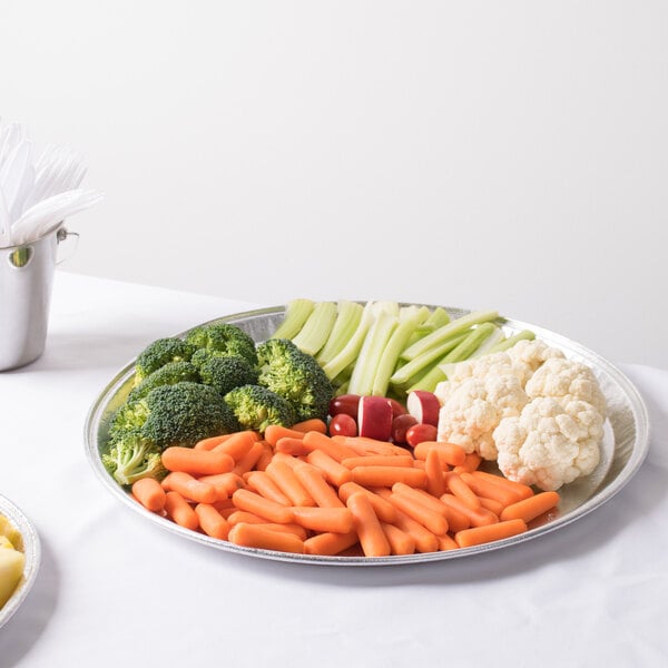 A Durable Packaging round foil catering tray with broccoli and carrots on a table.