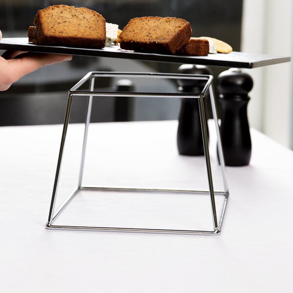 A hand holding a plate of bread on an American Metalcraft stainless steel square display stand.