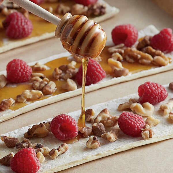 A white honey dipper drizzling Monarch's Choice Baker's Special Honey onto a piece of bread with nuts and raspberries.