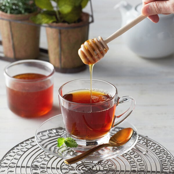 A hand using a Monarch's Choice honey dipper to drizzle honey into a glass cup of tea.