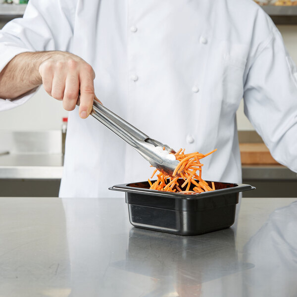 A chef using tongs to put carrots into a black Cambro food pan.