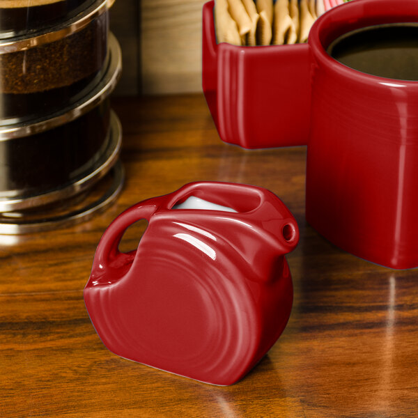 A red Fiesta mini disc creamer pitcher on a wood surface with a cup of coffee.