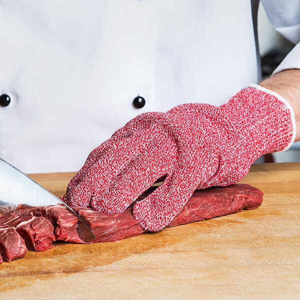 A person wearing a San Jamar red cut resistant glove cuts meat on a table.