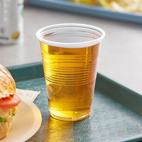 A translucent plastic cup of beer next to a tomato sandwich on a tray.