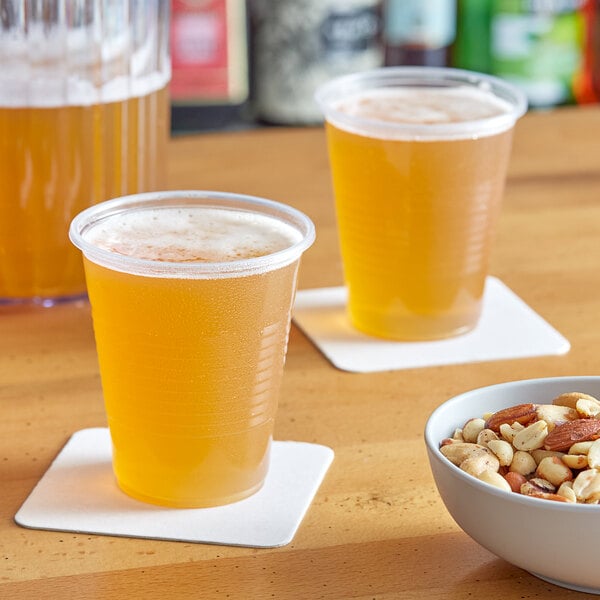 A close-up of a translucent plastic cup filled with yellow liquid on a table with a couple of cups of beer and a bowl of mixed nuts.