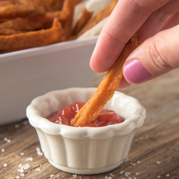 A person holding a Carlisle ivory scalloped ramekin filled with ketchup and dipping a french fry into it.