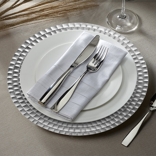 A white plate with a silver tiled charger plate on it with silverware and a white napkin.