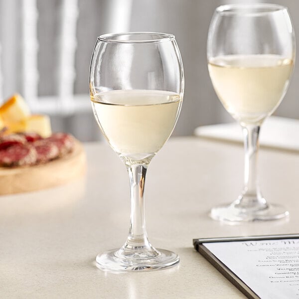 Two Acopa wine glasses on a table with a glass of white wine.