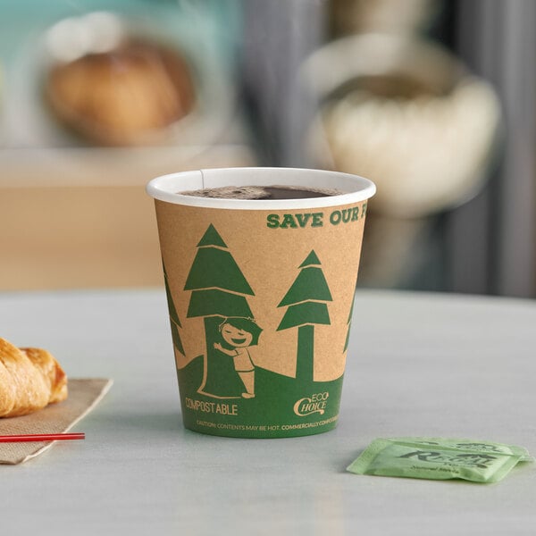 A close up of an EcoChoice Kraft Tree Print Paper Hot Cup full of coffee on a table with a croissant bag.