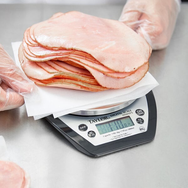 A gloved hand weighing a stack of sliced ham on a Taylor digital portion scale.