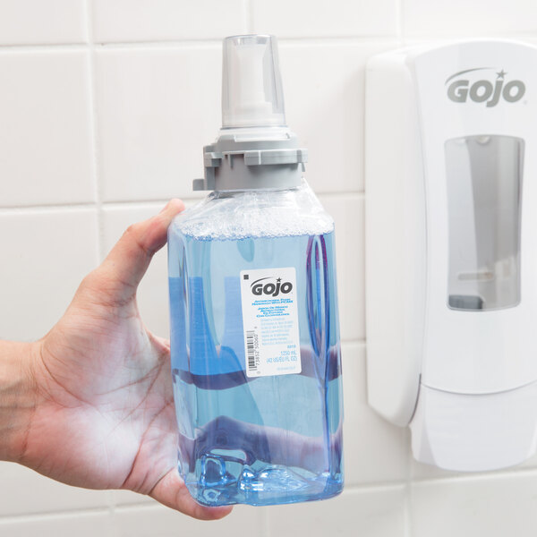 A hand holding a plastic container of GOJO foaming antimicrobial hand soap with blue liquid.