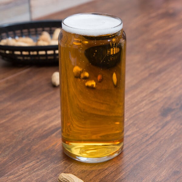 A Libbey can glass of beer on a table with peanuts.