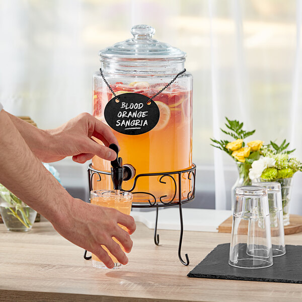 A person pouring a clear drink from an Acopa glass beverage dispenser with a metal stand into a glass.