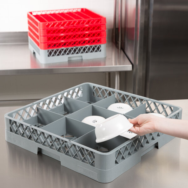 A hand placing a white bowl in a gray Noble Products glass rack with 9 compartments.