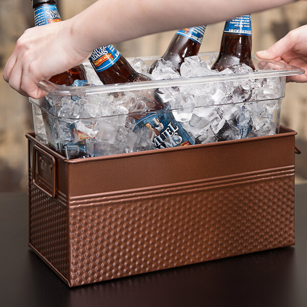 A person's hand reaching for a bottle of beer in a copper metal ice display filled with ice cubes.