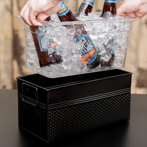 A hand holding a bottle of beer in a black metal ice display with ice and a clear food pan.