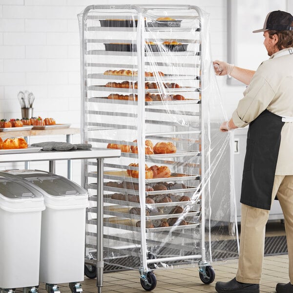 A man in a kitchen using a Regency clear plastic bun pan rack cover to transport pastries.