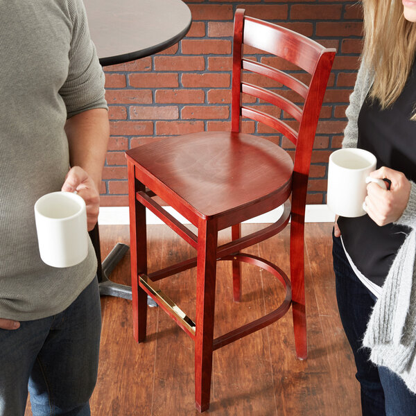 A man and woman holding coffee cups in front of a Lancaster Table & Seating mahogany wood ladder back bar stool with a mahogany wood seat.