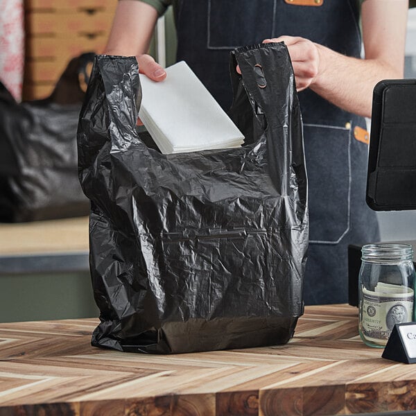 A person holding a black Choice medium-duty plastic T-shirt bag with white napkins inside.