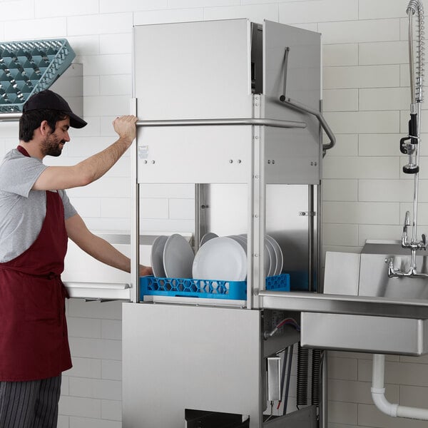 A man in a red apron using a Noble Warewashing tall door type dishwasher to wash plates in a professional kitchen.