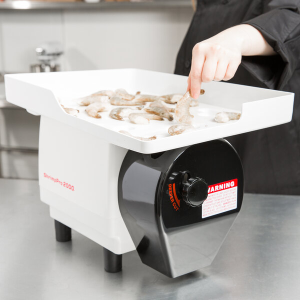 A hand using a Nemco ShrimpPro 2000 to cut and devein shrimp on a white tray.