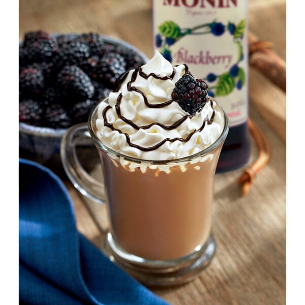 A glass cup of coffee with whipped cream and Monin blackberry syrup garnished with blackberries.