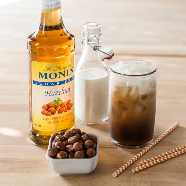 A bottle of Monin Sugar Free Hazelnut Flavoring Syrup next to a bowl of nuts and a glass of milk.