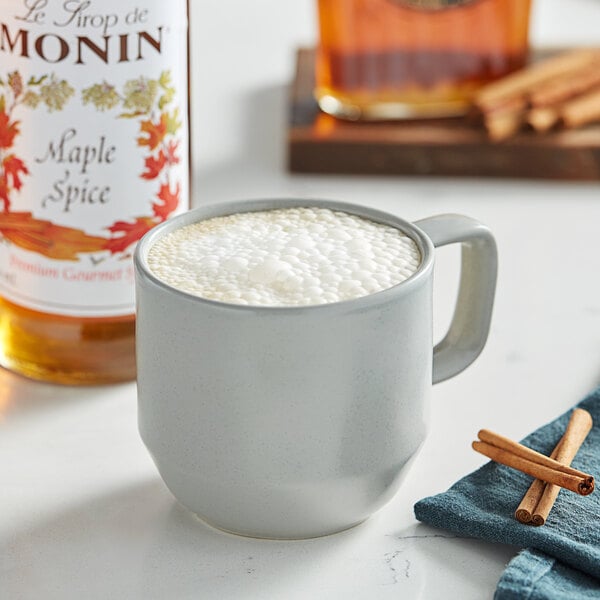 A mug of hot drink with foam and cinnamon sticks next to a bottle of Monin Premium Maple Spice Flavoring Syrup.