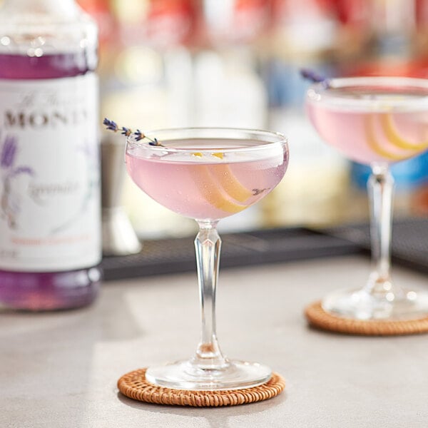 Two glasses of pink lavender cocktails with lemons and lavender on a table with a bottle of Monin Premium Lavender Flavoring Syrup.