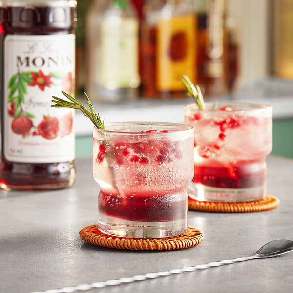 Two glasses of red liquid with Monin Pomegranate Syrup garnish on a table.