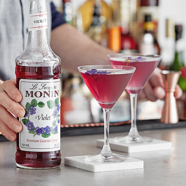 A person holding a bottle of Monin violet flavoring syrup next to glasses of pink drinks.
