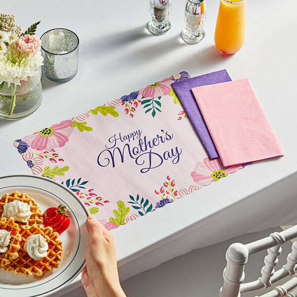 A woman's hand sets a Mother's Day placemat on a table with waffles, whipped cream, and strawberries.