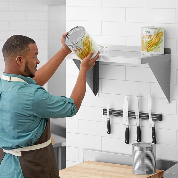 A man in a professional kitchen wearing an apron and holding a can of corn on a Regency stainless steel wall shelf.