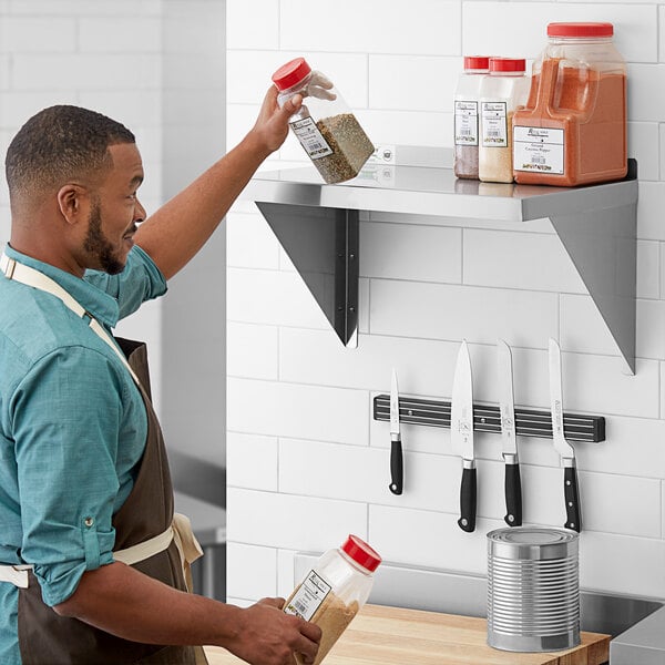 A man in a brown apron and blue shirt holding a bottle of brown powder over a stainless steel shelf.
