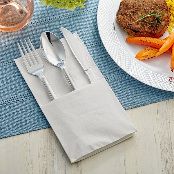 A Visions flared heavy weight silver plastic fork and knife in a white napkin next to a plate of food.