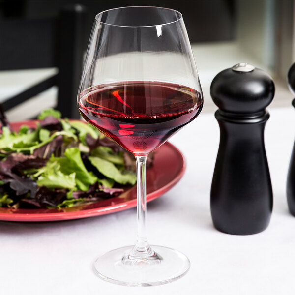 A Spiegelau burgundy wine glass filled with red wine next to a plate of salad.