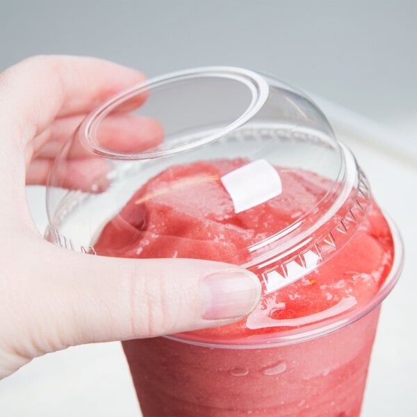A hand holding a Dart clear plastic cup with a red smoothie inside and a clear plastic dome lid with a hole in it.