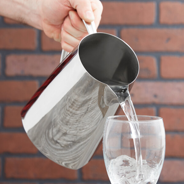 A hand pouring water from a Vollrath Triennium stainless steel water pitcher into a glass.