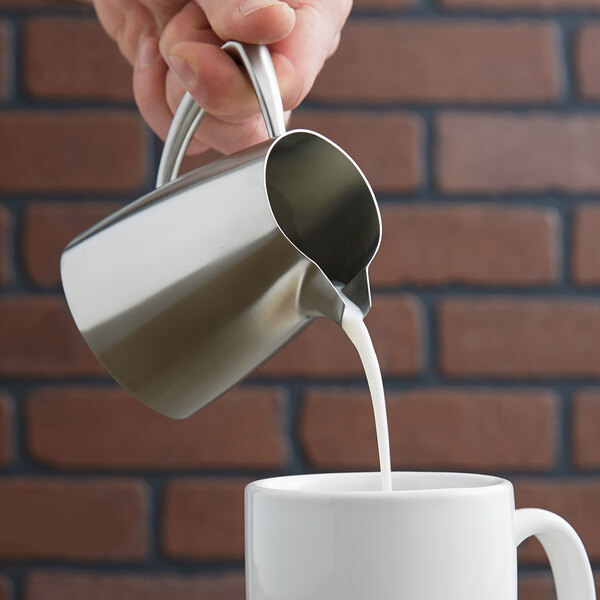 A hand pouring milk into a Vollrath stainless steel creamer.