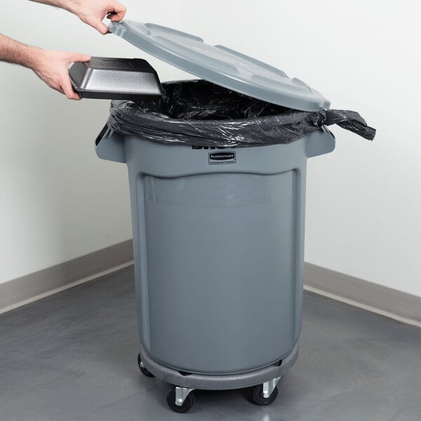 A person using a tablet to put a plastic lid on a Rubbermaid trash can.