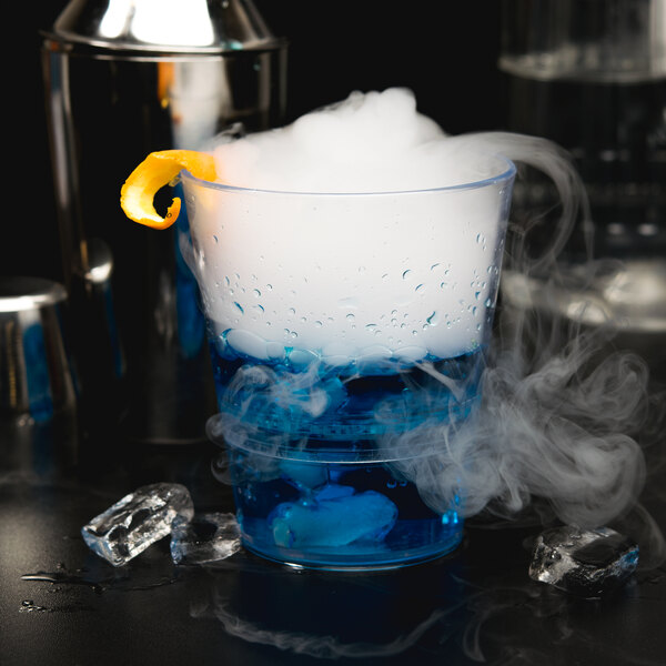 A clear WNA Comet plastic rocks glass filled with a blue drink with ice and smoke, garnished with a slice of orange.