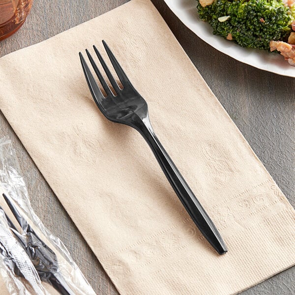 A black plastic Choice fork on a napkin next to a plate of food.