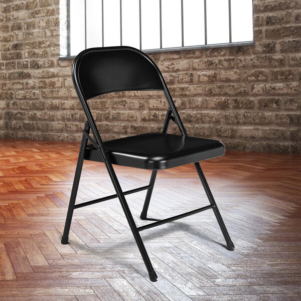 A National Public Seating black metal folding chair in front of a brick wall.