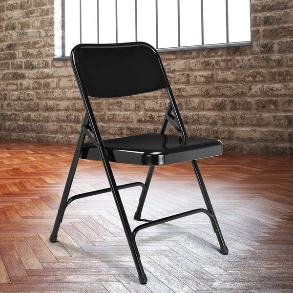A National Public Seating black metal folding chair in front of a brick wall.