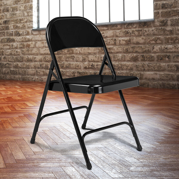 A black National Public Seating metal folding chair in front of a brick wall.