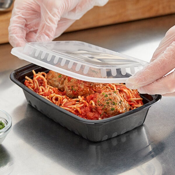 A person in gloves holding a black rectangular plastic container with a plastic lid filled with spaghetti.