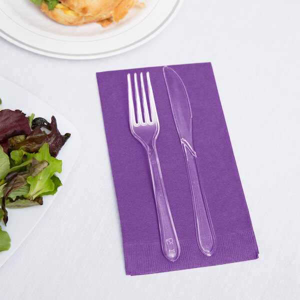A purple Creative Converting guest towel with a fork and knife on it next to a plate of salad.